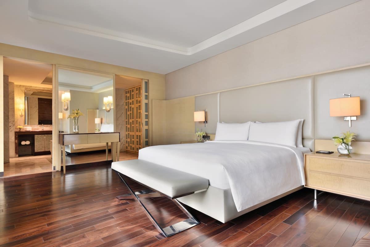 A modern hotel room with a large bed, a bench, a bedside table with a lamp, and a mirror.