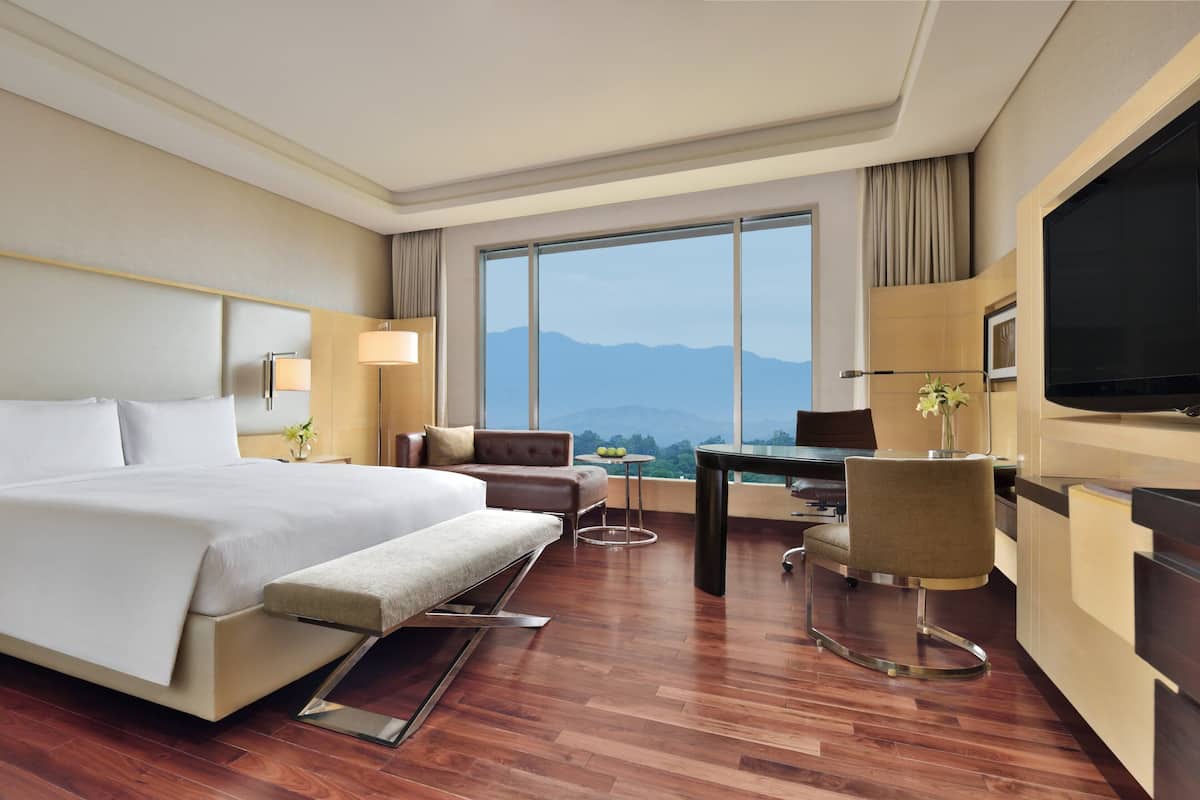A hotel room with a large bed, a desk, a chair, a TV, and a view of mountains through a large window.