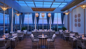 Un área de comedor en la azotea con mesas, sillas, ventanas amplias y luces colgantes.