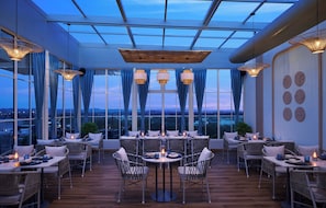 Un área de comedor en la azotea con mesas, sillas, ventanas amplias y luces colgantes.