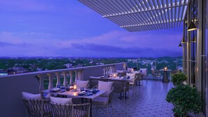 Una terraza en la azotea con mesas y sillas, vista panorámica de la ciudad y una estructura de pérgola.