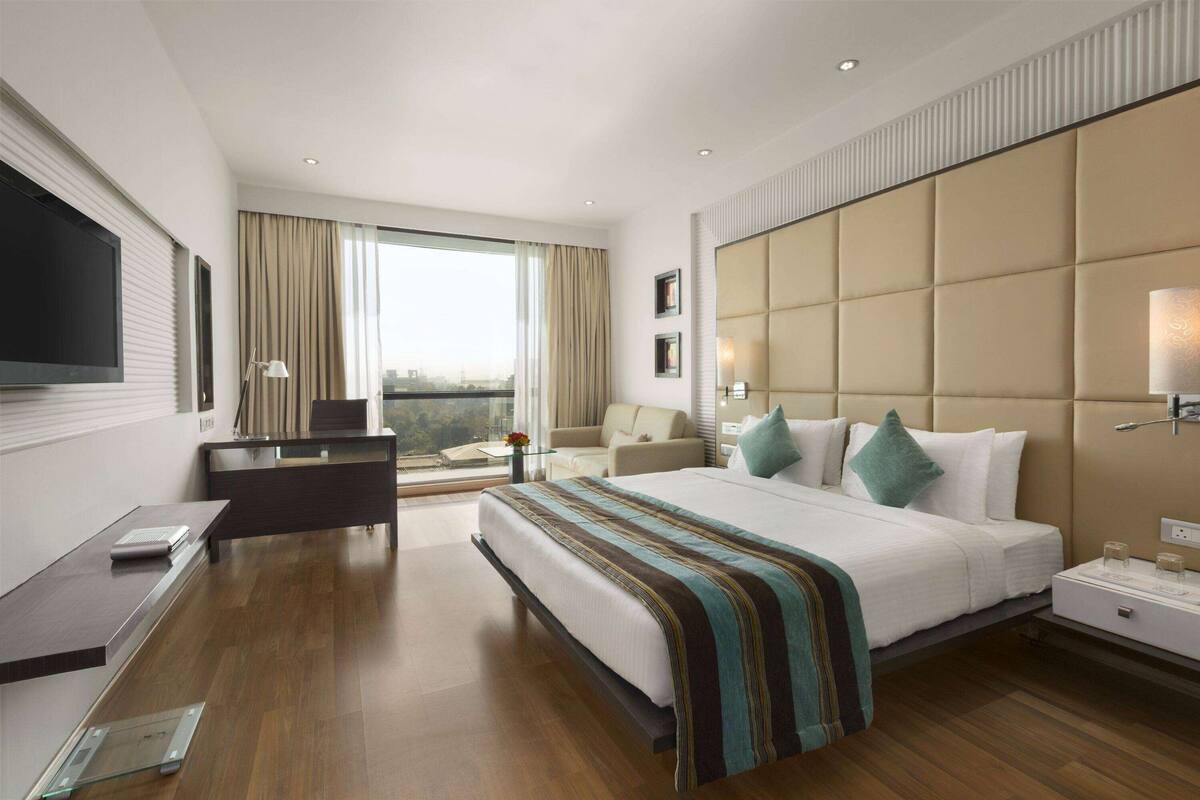 A modern hotel room with a large bed, a desk, a chair, a TV, and a window with a city view.