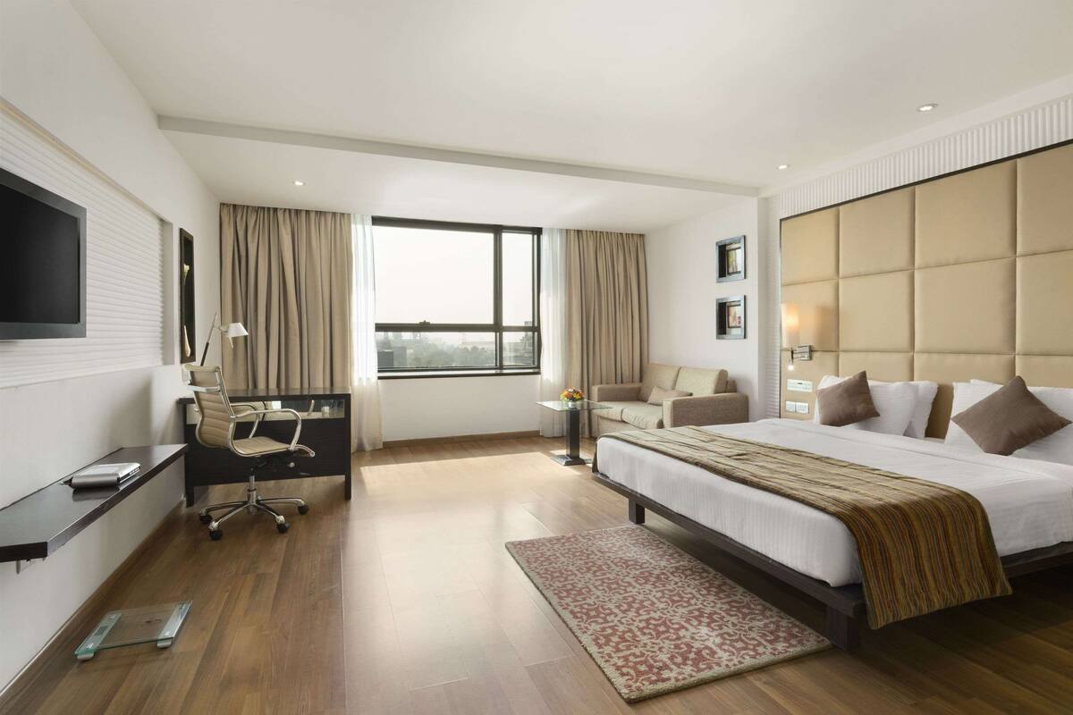 A modern hotel room with a large bed, a desk with a chair, a flat-screen TV, and a large window with curtains.