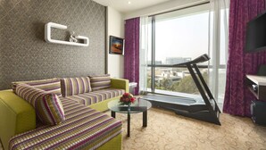 A modern living room with a sofa, a coffee table with a vase, a treadmill, and a large window with curtains. - Ramada by Wyndham Navi Mumbai (Navi Mumbai)