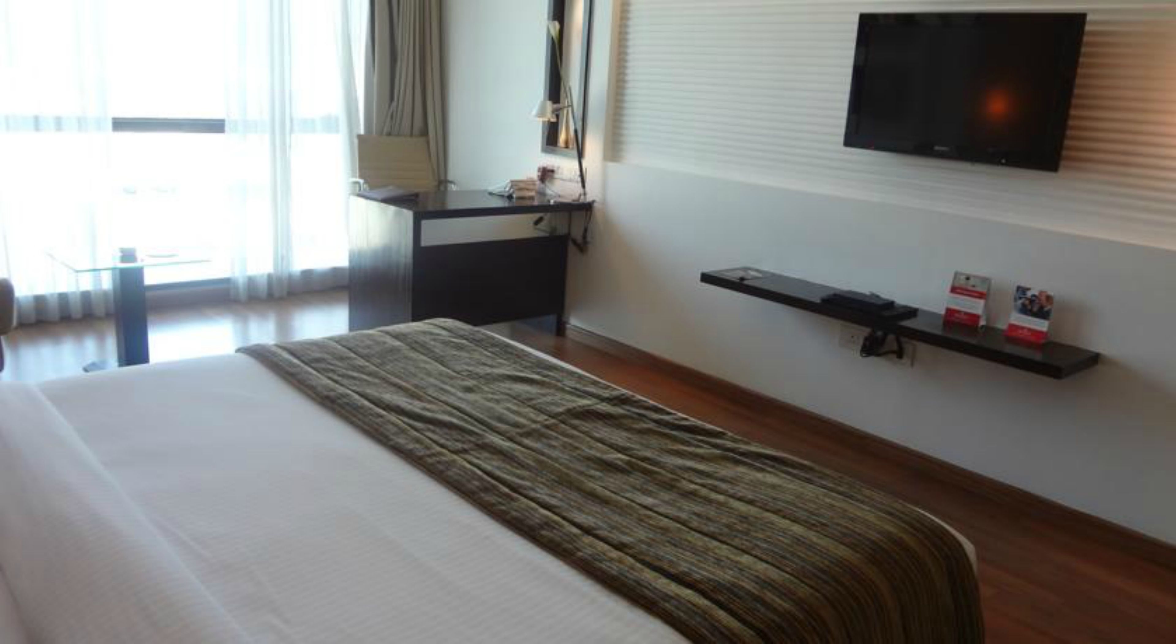 Habitación de hotel con cama, escritorio, televisor montado en la pared y ventana con cortinas.