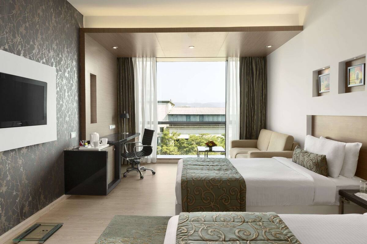 A hotel room with two beds, a desk, a chair, a TV, and a balcony with a view.
