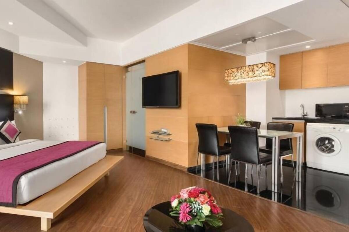 A modern hotel room with a bed, a desk, a chair, a TV, a washing machine, and a small table with flowers.
