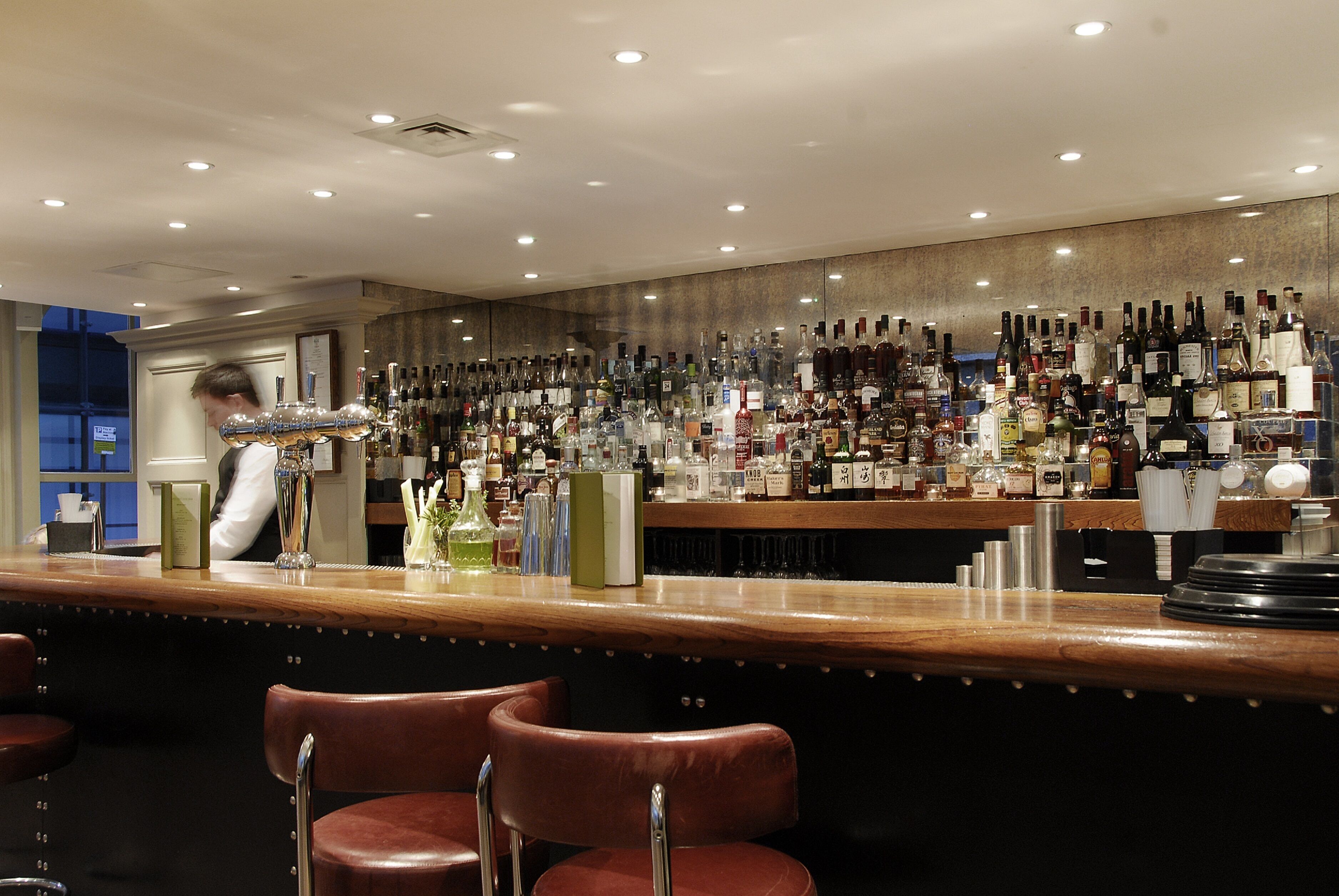 Un espace bar avec un comptoir en bois, de nombreuses bouteilles d’alcool et deux tabourets de bar en cuir.