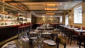 A restaurant with multiple dining tables set for guests, a bar area with shelves stocked with bottles, and a brick wall with framed pictures. - Montcalm Brewery, London City (London)