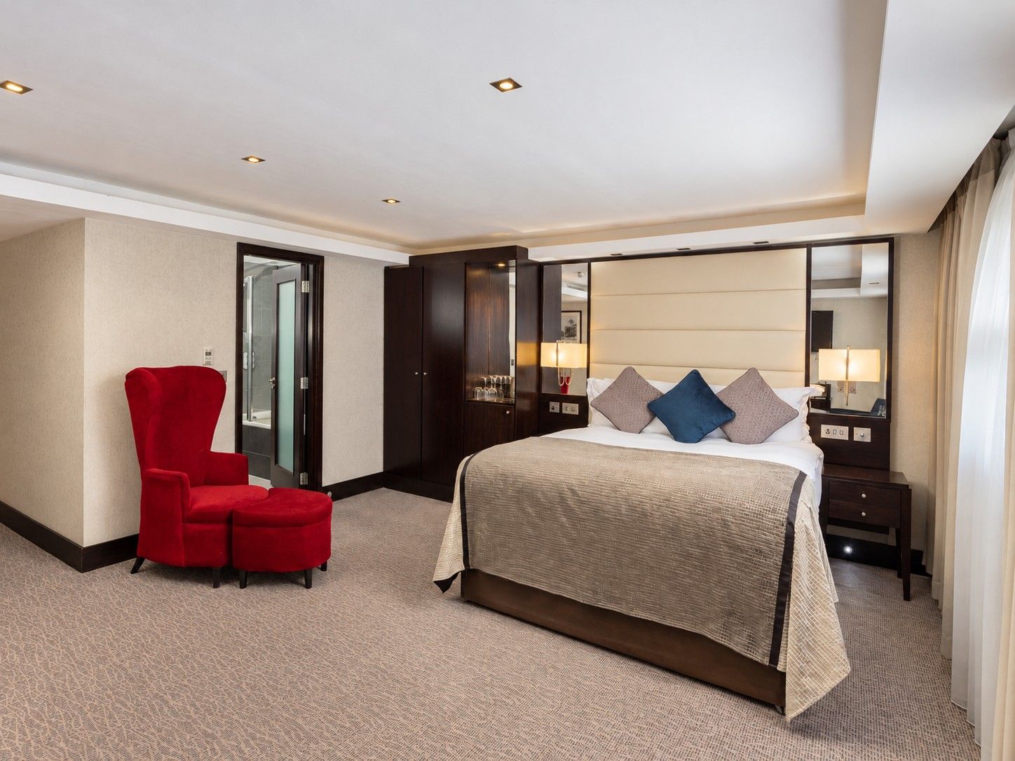 A hotel room with a bed, a red armchair, a wardrobe, a mirror, and a bedside table with a lamp.