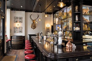 Un espace bar avec un comptoir en bois, des tabourets rouges, une tête de cerf accrochée au mur et un présentoir à vin.