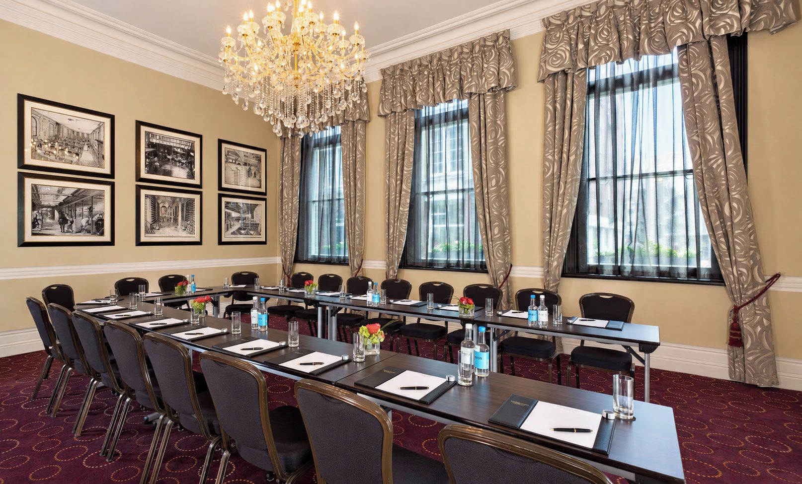 A conference room with a long table, chairs, a chandelier, framed pictures on the wall, and large windows with curtains.