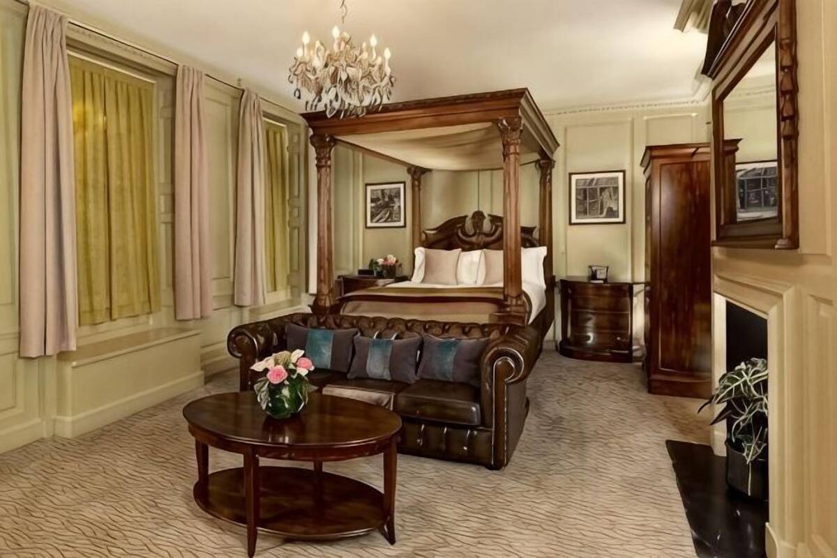 Een kamer met een hemelbed, een bank, een open haard en een kroonluchter, met een kamerbreed tapijt en gordijnen.