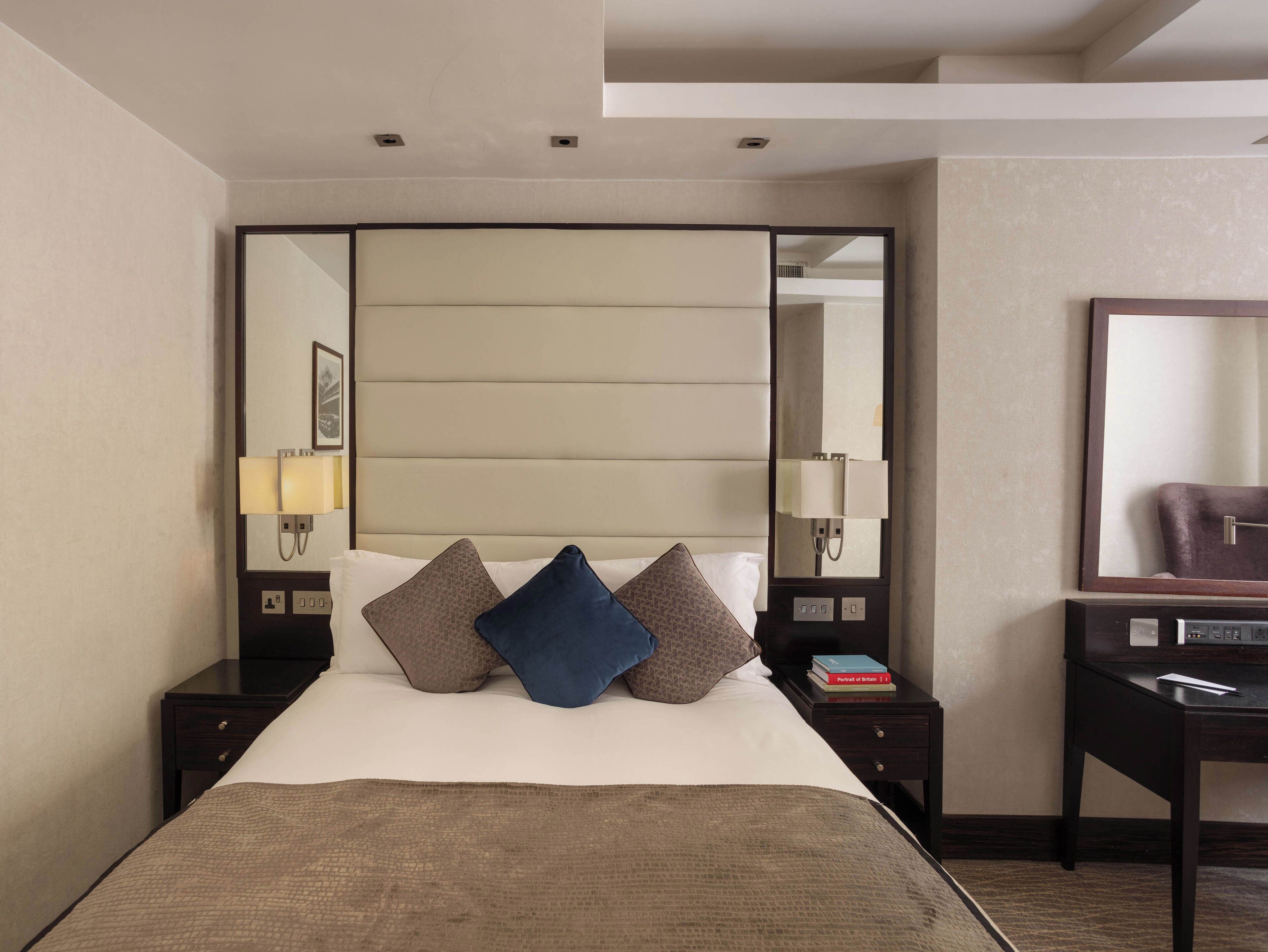Habitación de hotel con una cama grande, cabecera con iluminación integrada, mesita de noche con lámpara y espejo en la pared.