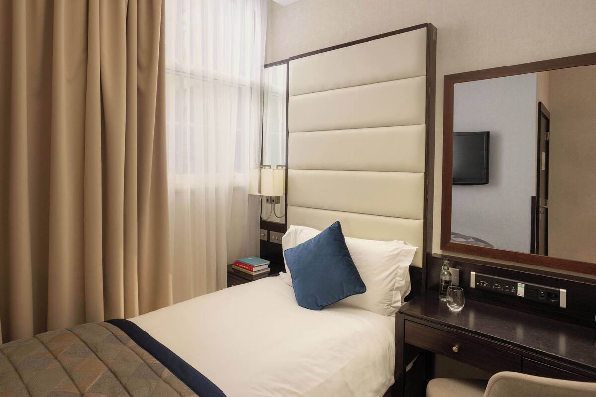 Een hotelkamer met een bed, nachtkastje, spiegel, televisie en raam met gordijnen.