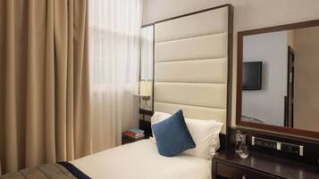 Una habitación de hotel con cama, mesita de noche, espejo, televisor y ventana con cortinas.