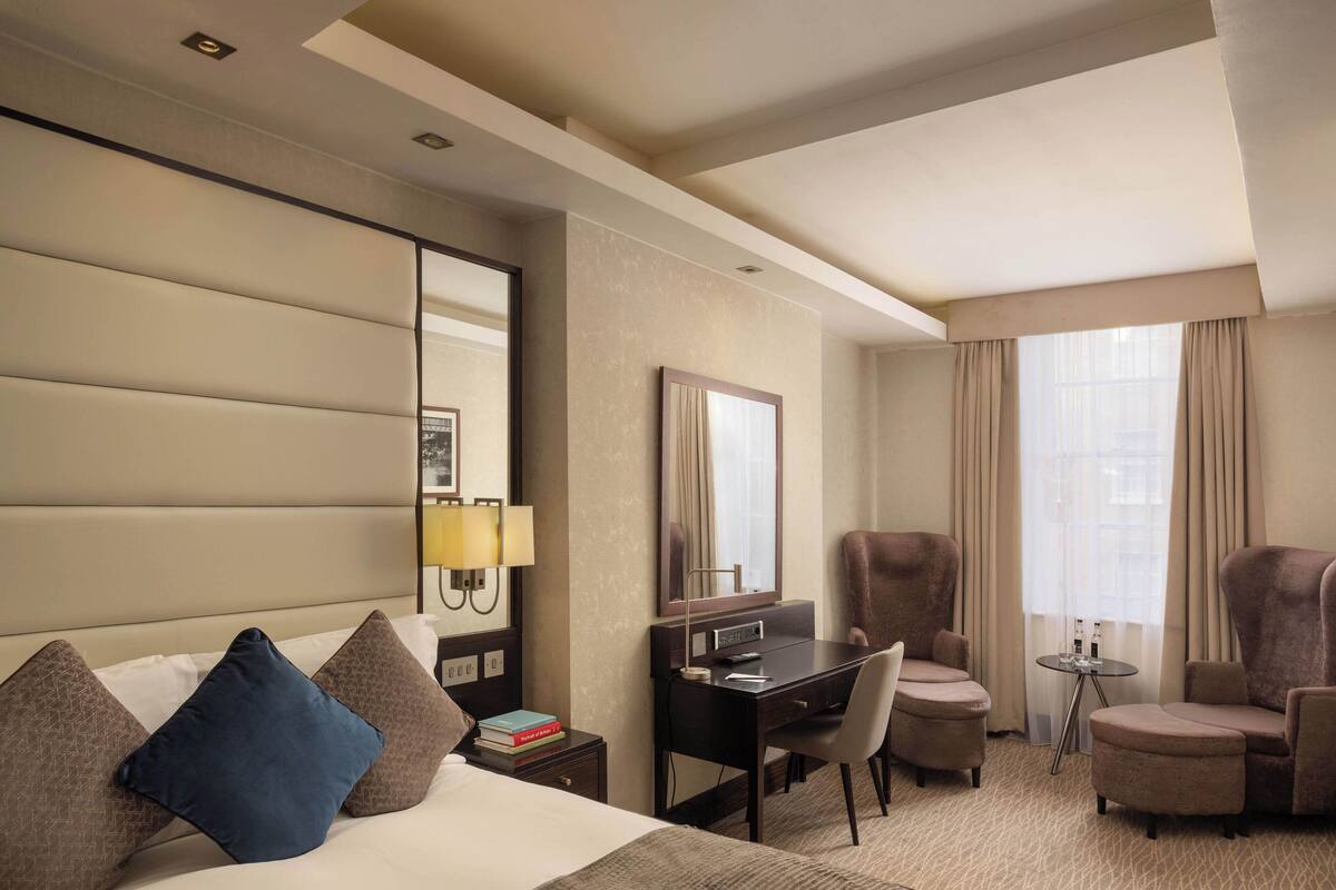 Een hotelkamer met een bed, een bureau, twee fauteuils, een kleine tafel en een groot raam met gordijnen.