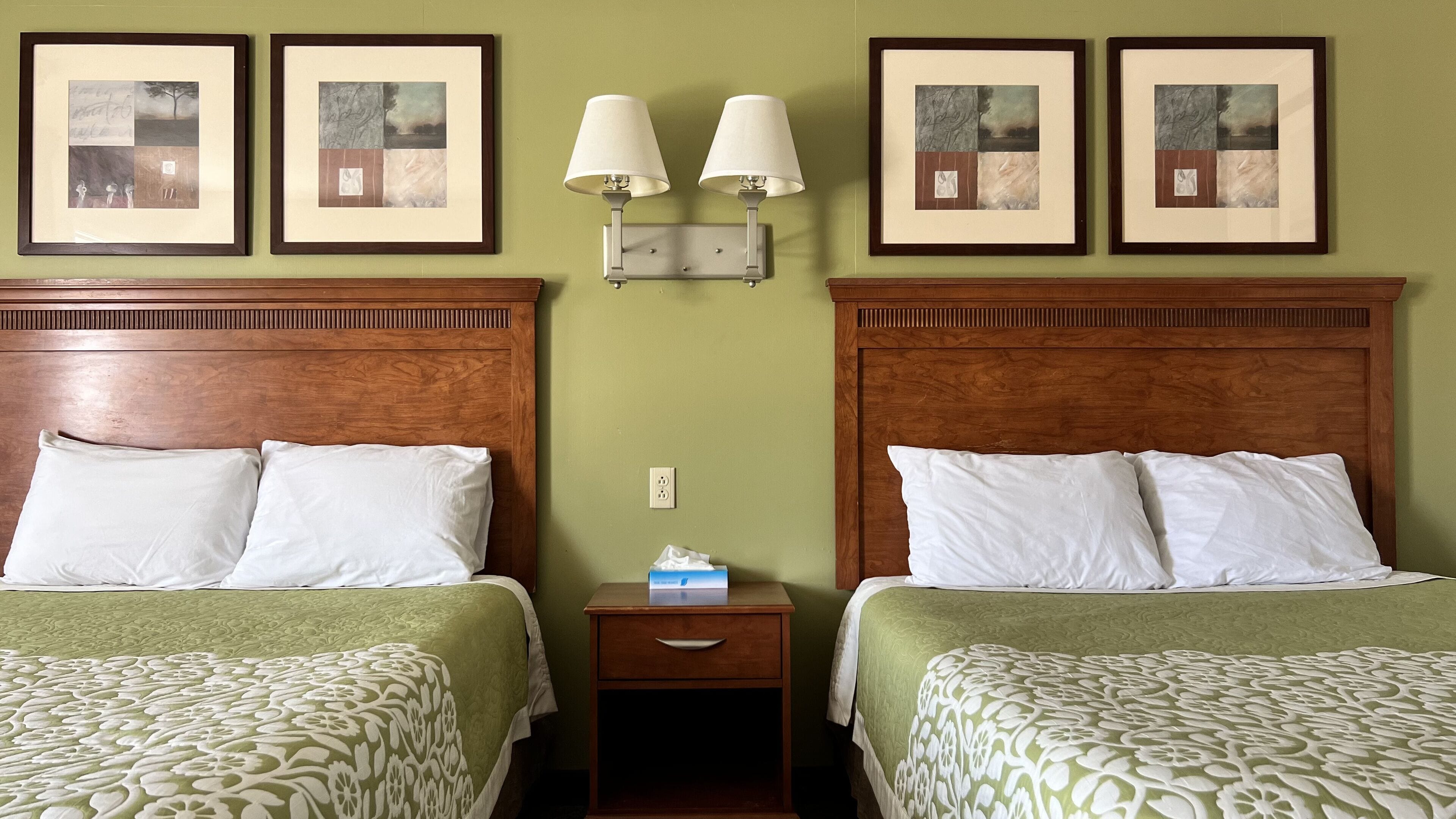 Une chambre d’hôtel avec deux lits, chacun doté d’une tête de lit en bois et recouvert d’une couvre-lit à motifs verts. Des tableaux encadrés sont accrochés au mur au-dessus des lits, et une table de chevet avec deux lampes fixées au mur se trouve entre les deux lits.