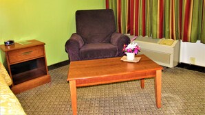Habitación de hotel con un sillón morado, una mesita de noche de madera, una pequeña mesa con un florero y un equipo de aire acondicionado grande frente a una pared verde con cortinas a rayas.