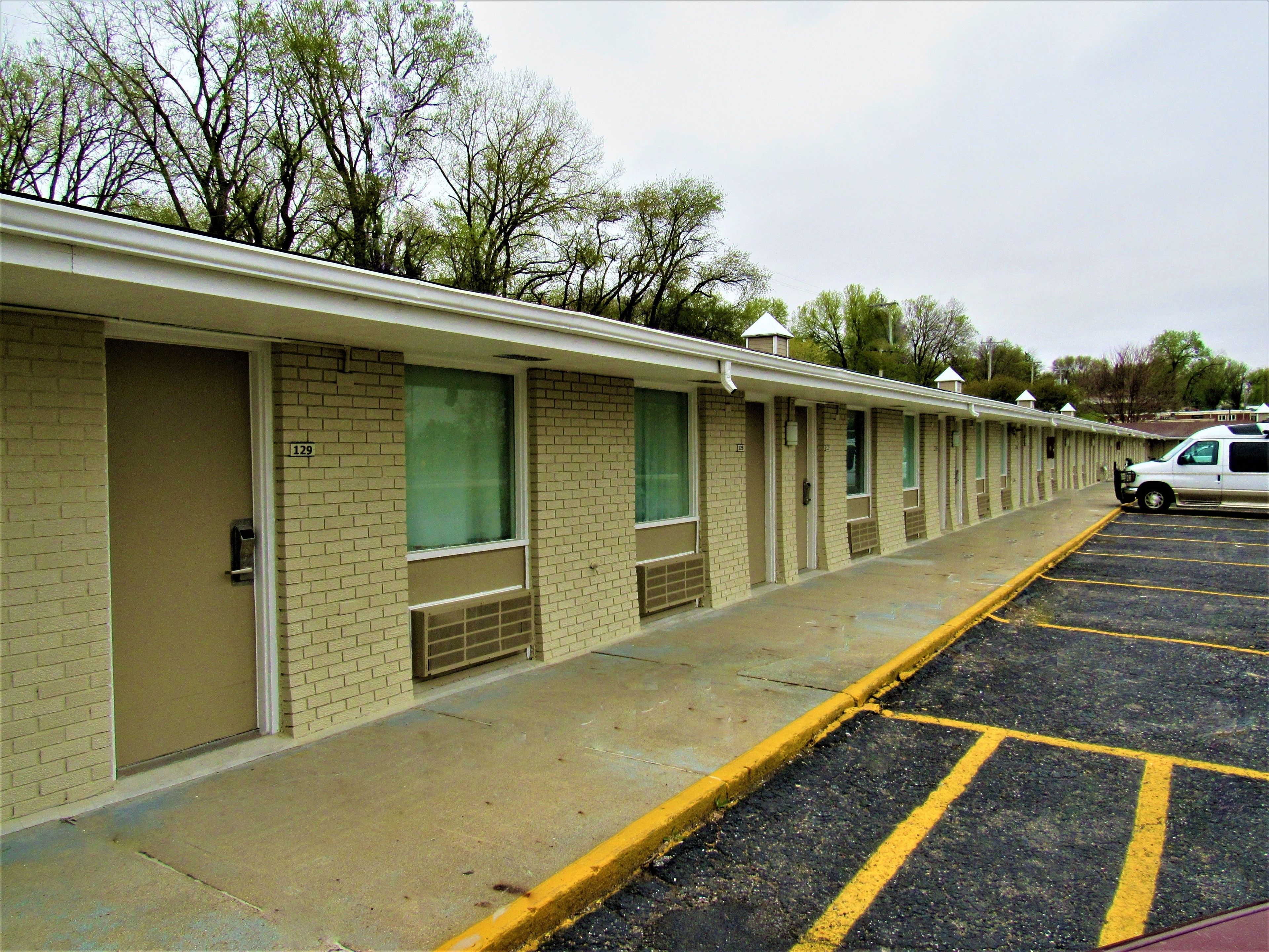 A row of motel rooms with individual doors, a parking lot, and a white van parked in the lot.
