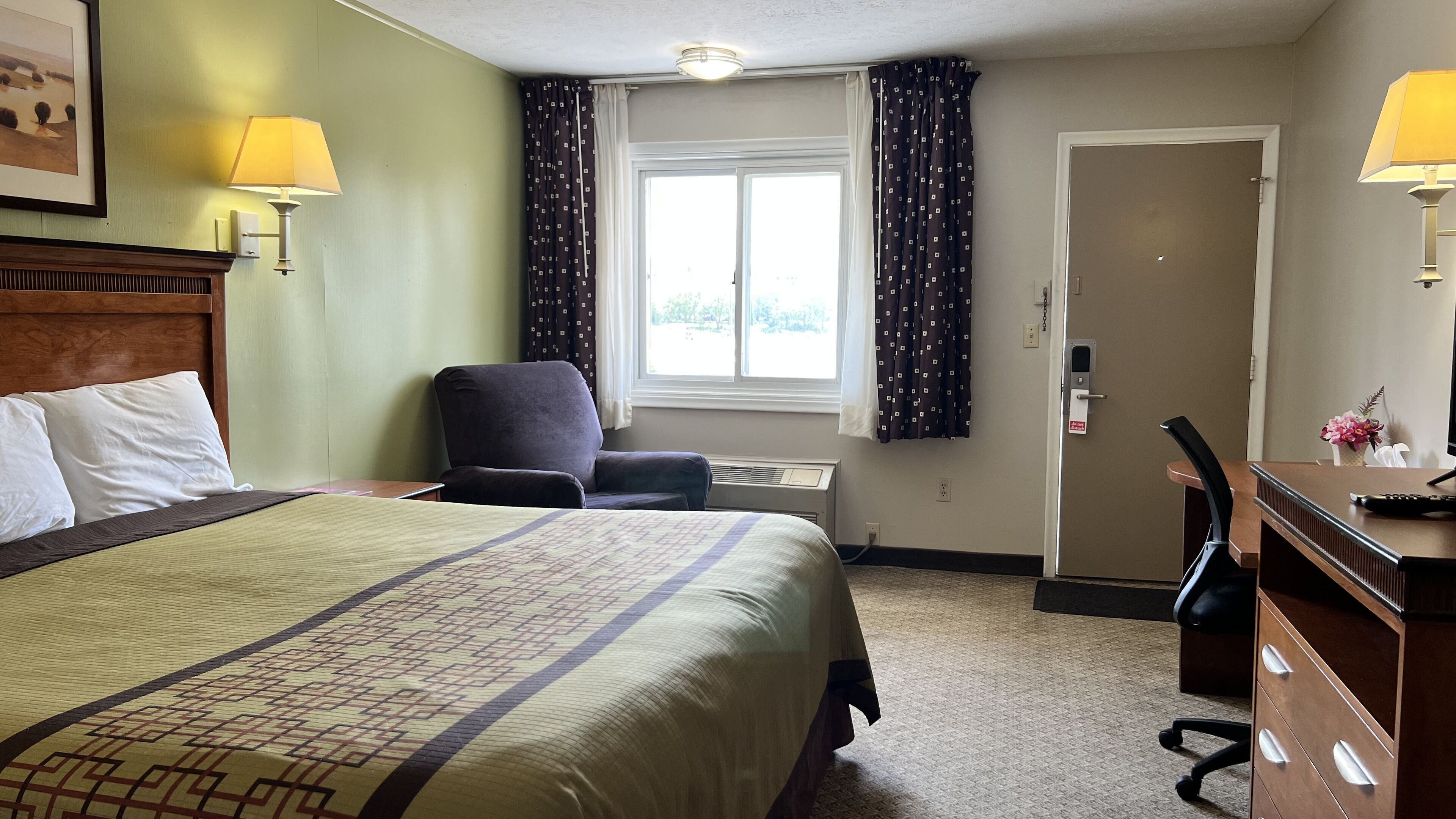 Une chambre d’hôtel avec un grand lit, un bureau, une chaise, une fenêtre avec des rideaux et une porte.