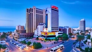 A cityscape with a prominent hotel featuring a large sign, multiple high-rise buildings, and a busy street with vehicles and palm trees. - Temeisi Hotel (Jieyang)