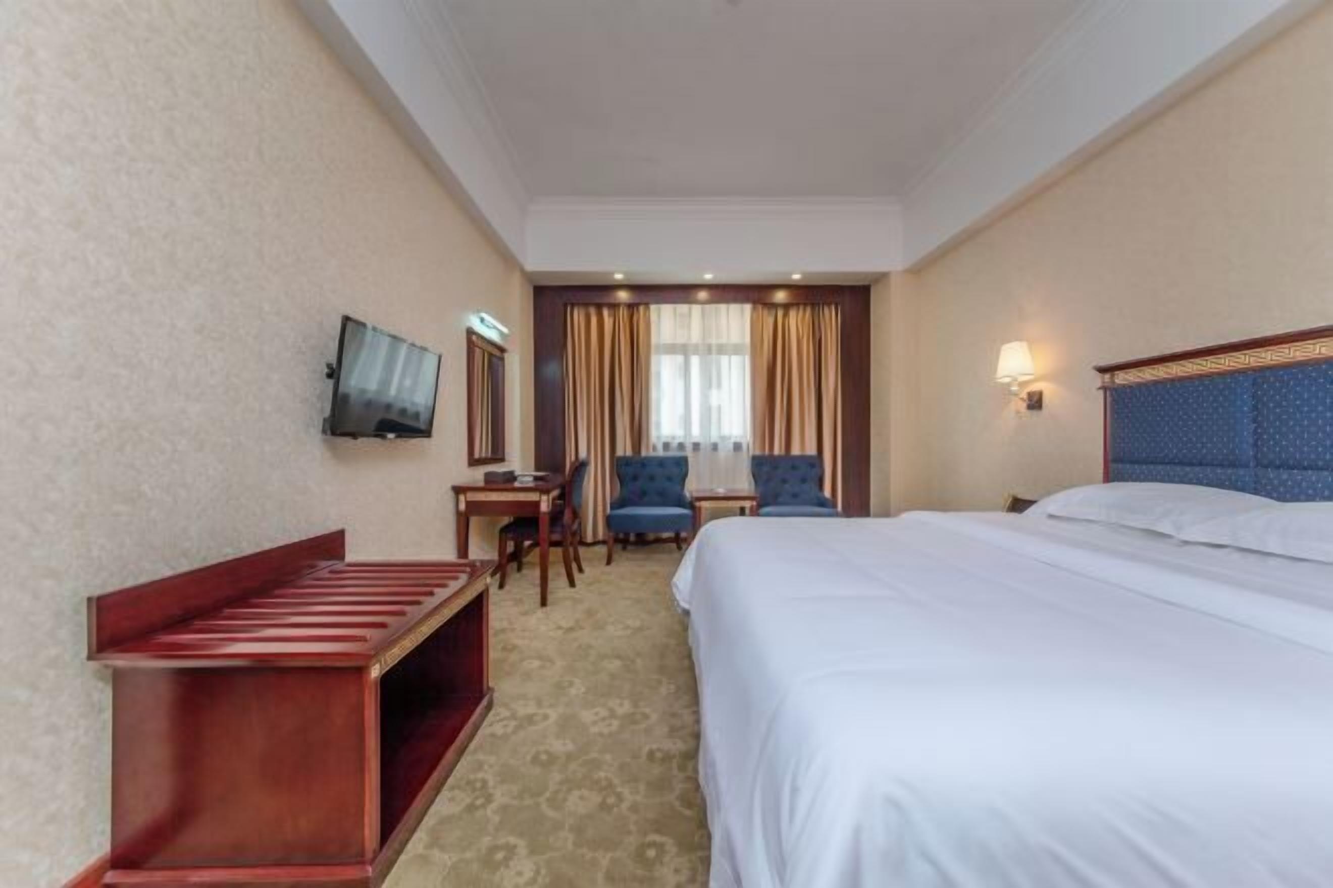 A hotel room with a large bed, a desk with a chair, a TV mounted on the wall, and a window with curtains.