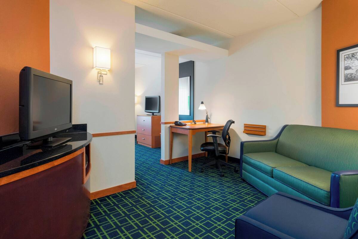 A hotel room with a television, desk, chair, sofa, and a large mirror.