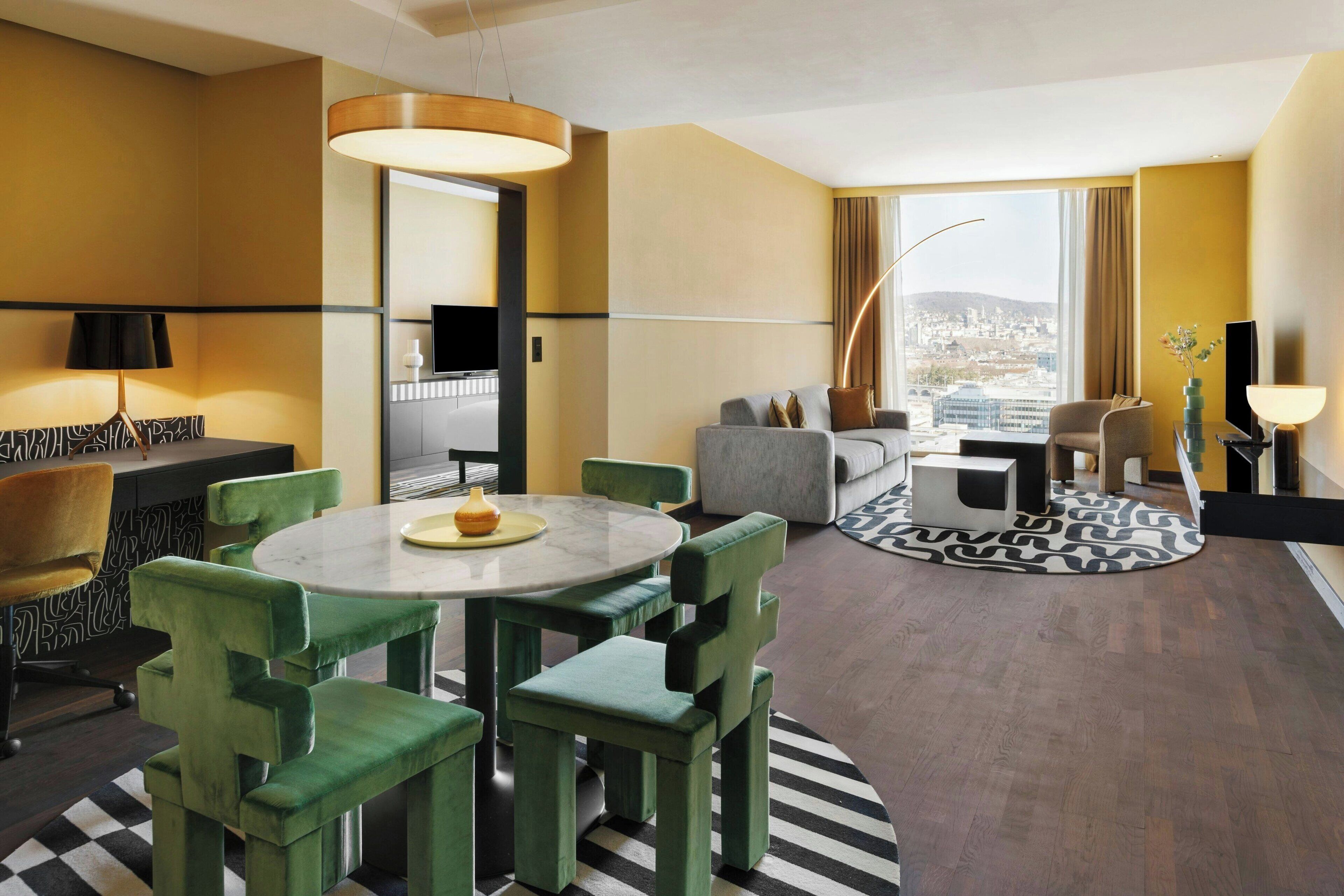 A hotel room with a dining area, a sofa, a TV, and a view of the city.