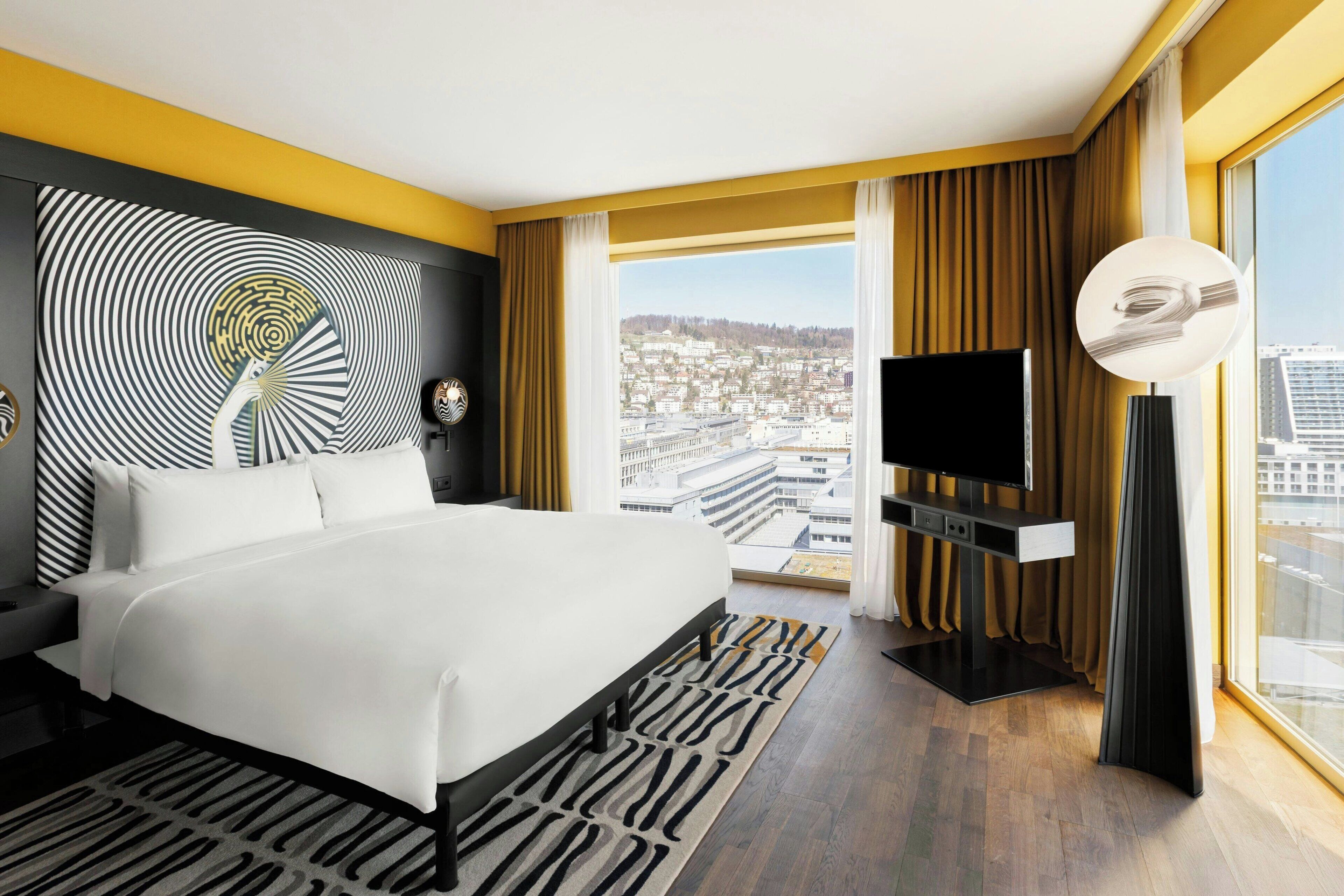 A modern hotel room with a large bed, a flat-screen TV, and a city view through large windows.