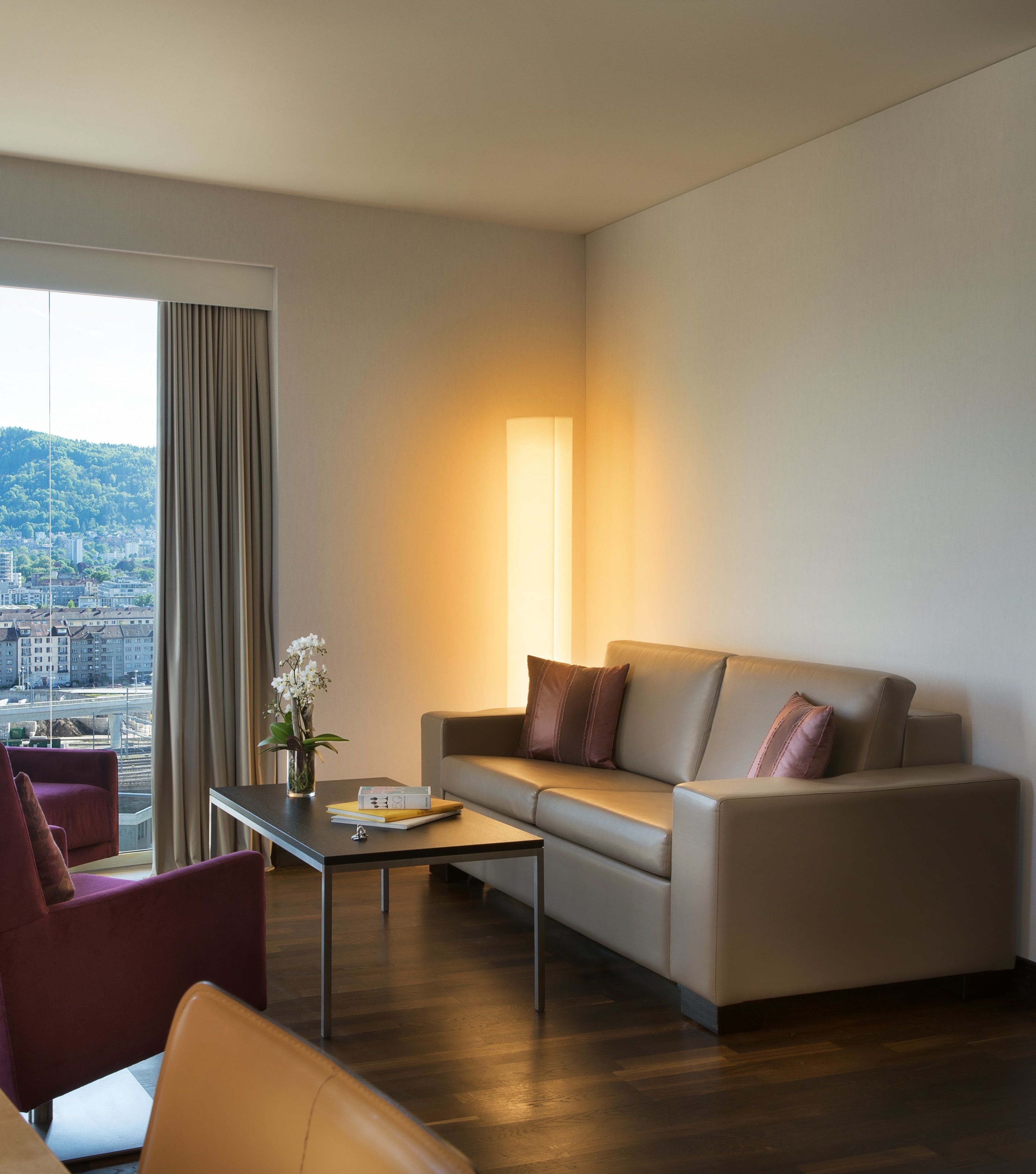 A modern hotel room with a sofa, armchairs, a coffee table, and a large window offering a city view.