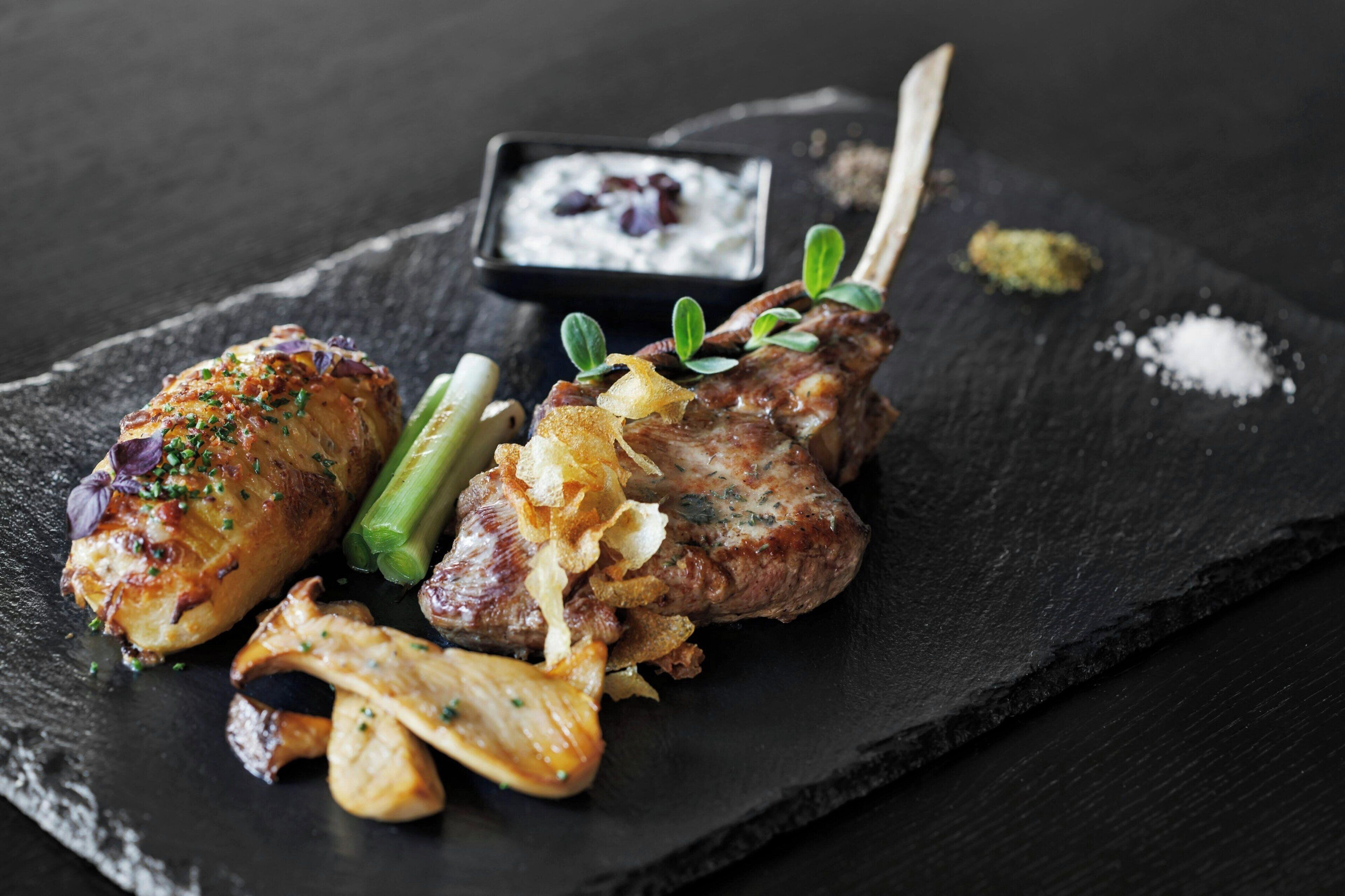 Une planche de service en ardoise noire, sur laquelle sont disposés de la viande grillée, des légumes et un petit bol de sauce.