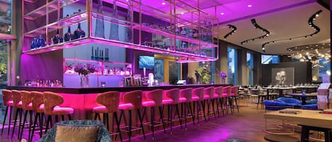Un espace bar moderne doté d’une étagère à plusieurs niveaux où sont exposés différents types de verres, d’un comptoir de bar avec un téléviseur et de sièges recouverts d’un tissu aux couleurs vives.