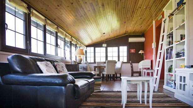 A spacious living room with a wooden ceiling, a black leather sofa, a white table, and a bookshelf.