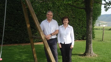 A man and a woman standing in a grassy area with a wooden swing set and a hedge in the background.