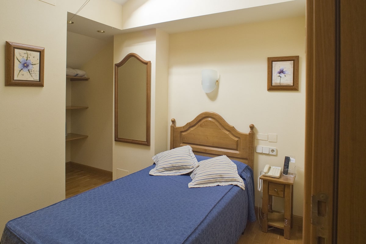 A bedroom with a skylight, a bed with a blue bedspread, a wooden headboard, a bedside table with a lamp, and a nightstand with a phone.