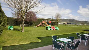 Un espacio al aire libre con césped, que incluye un parque infantil con toboganes y estructuras para escalar, además de un patio con mesas y sillas.