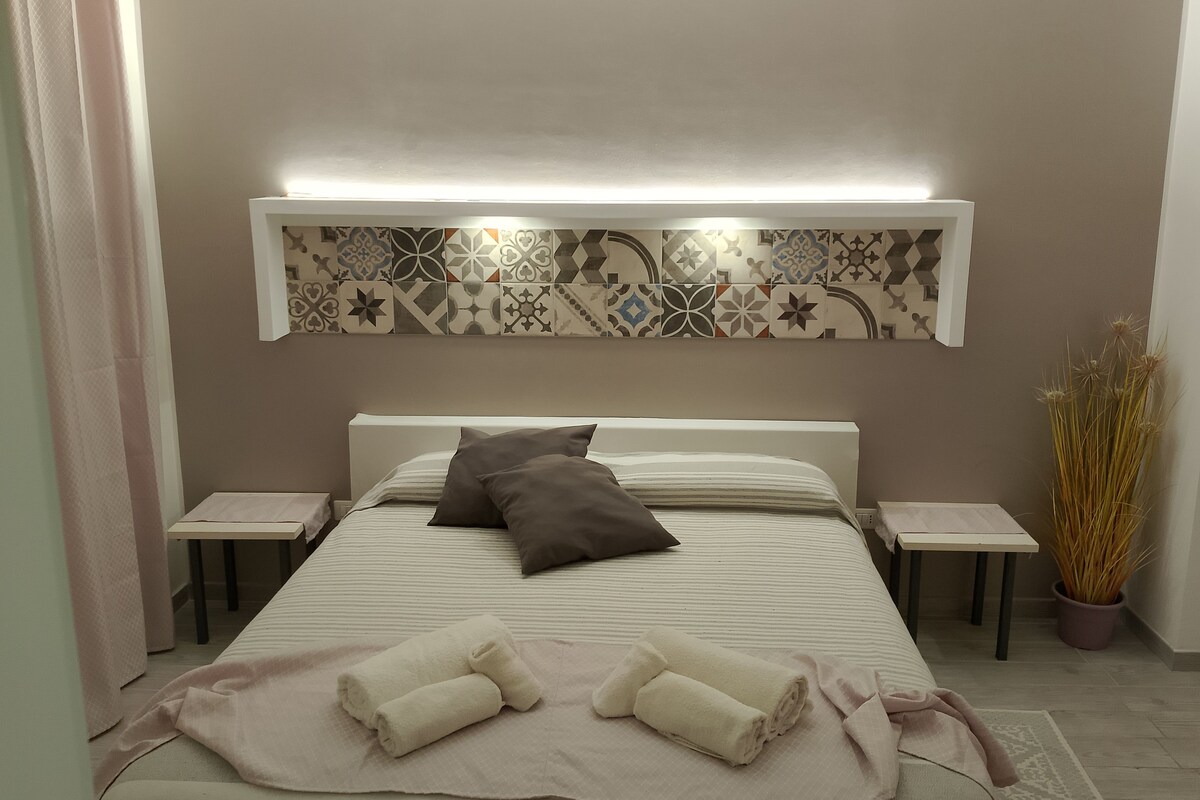 A neatly made bed with pillows, a bedside table, a decorative wall panel, and a potted plant.