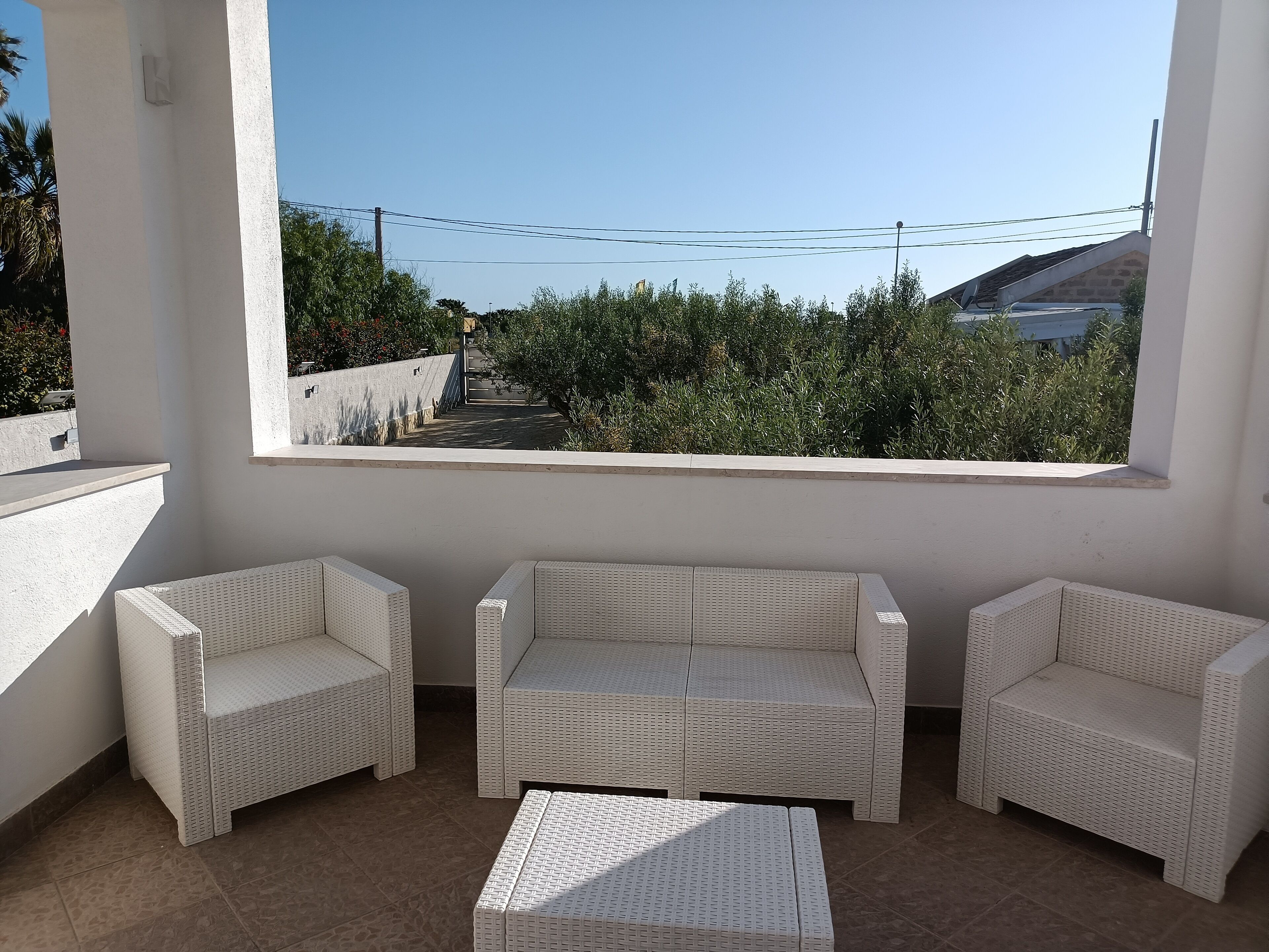 A balcony with white wicker furniture, including two chairs and a sofa, and a view of greenery and buildings.