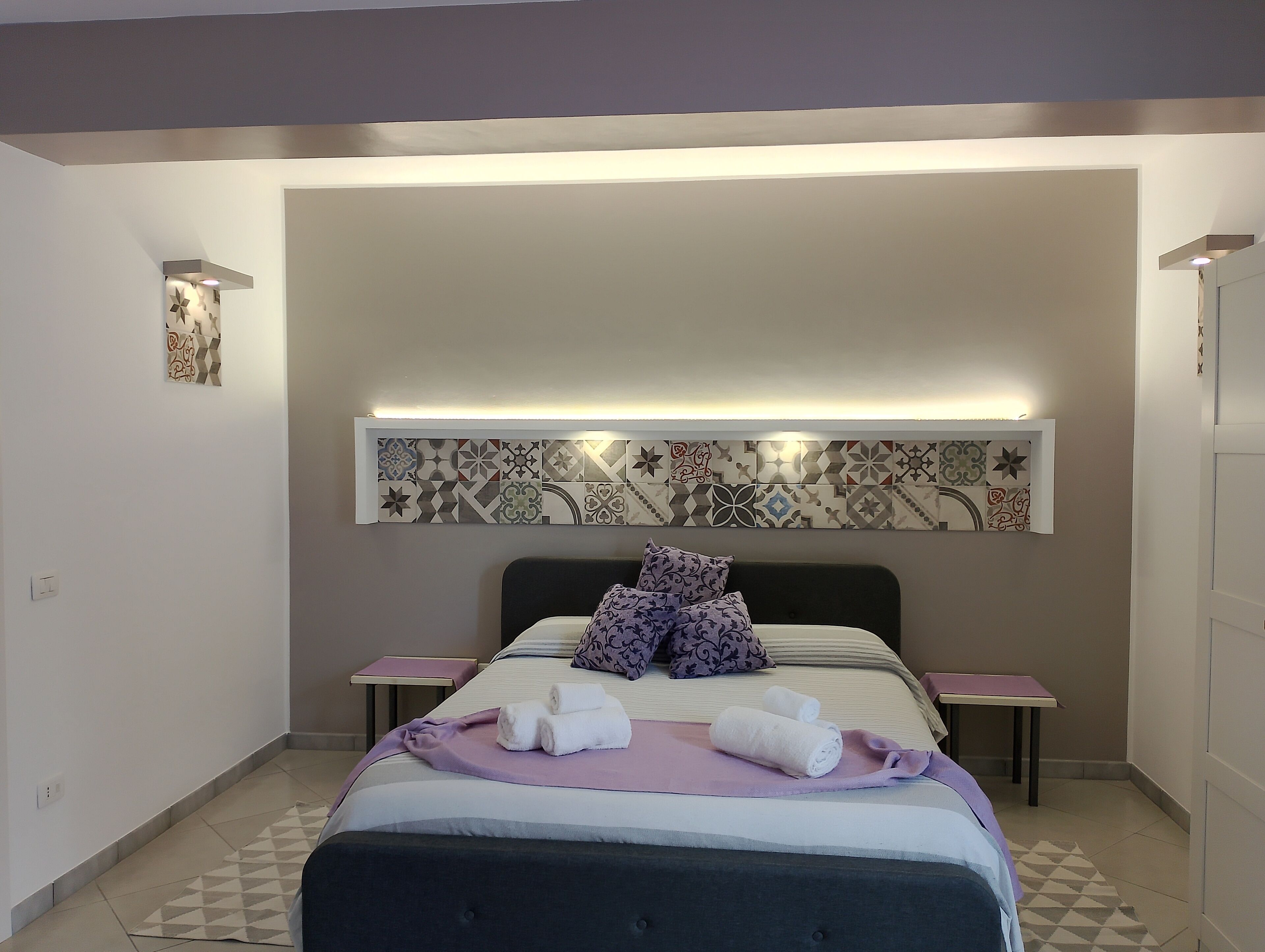 Een moderne slaapkamer met een bed, twee nachtkastjes, een wandlamp en een decoratieve wand van tegels.