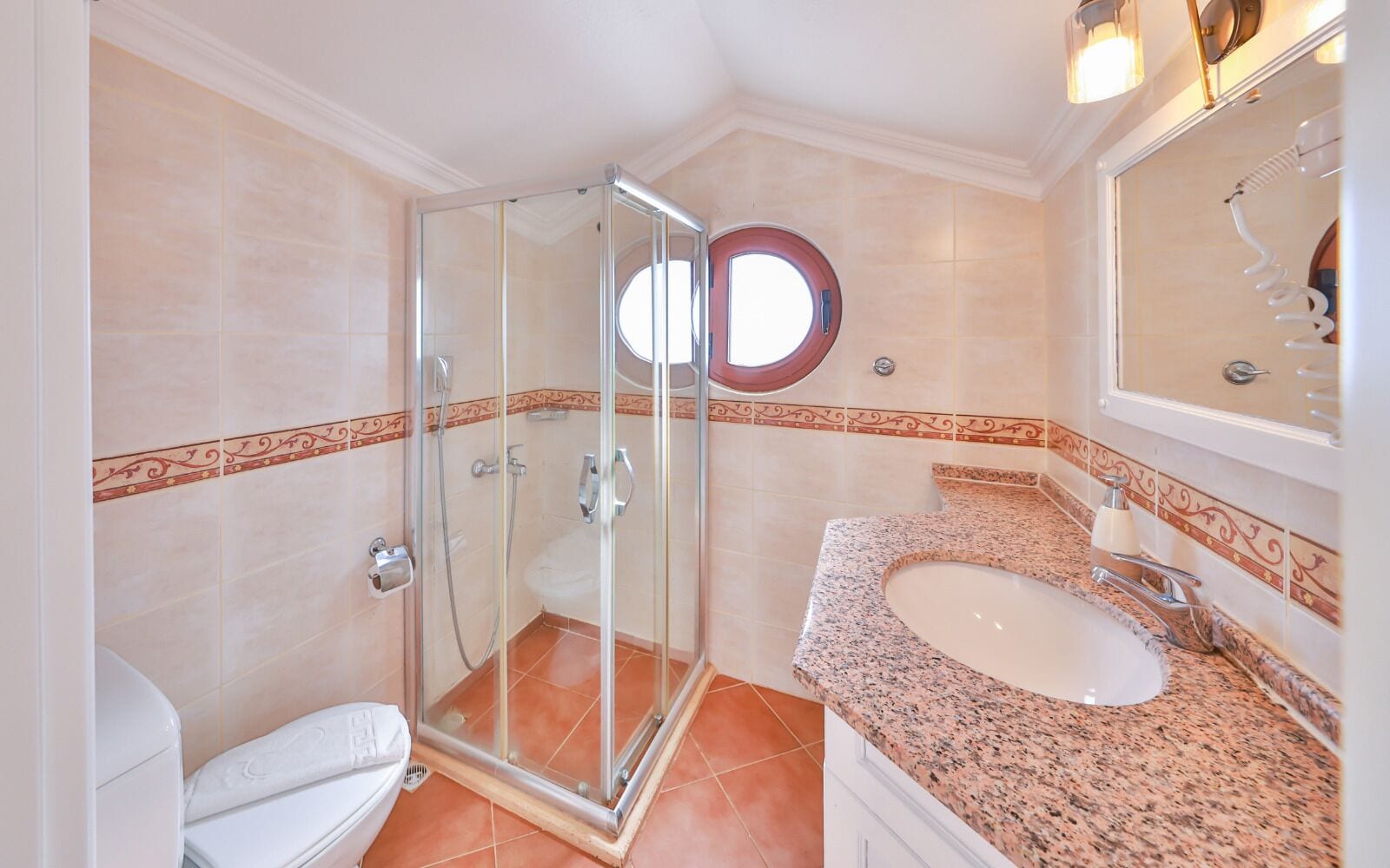 Un baño con ducha envuelta en vidrio, inodoro blanco y lavamanos sobre encimera de granito.