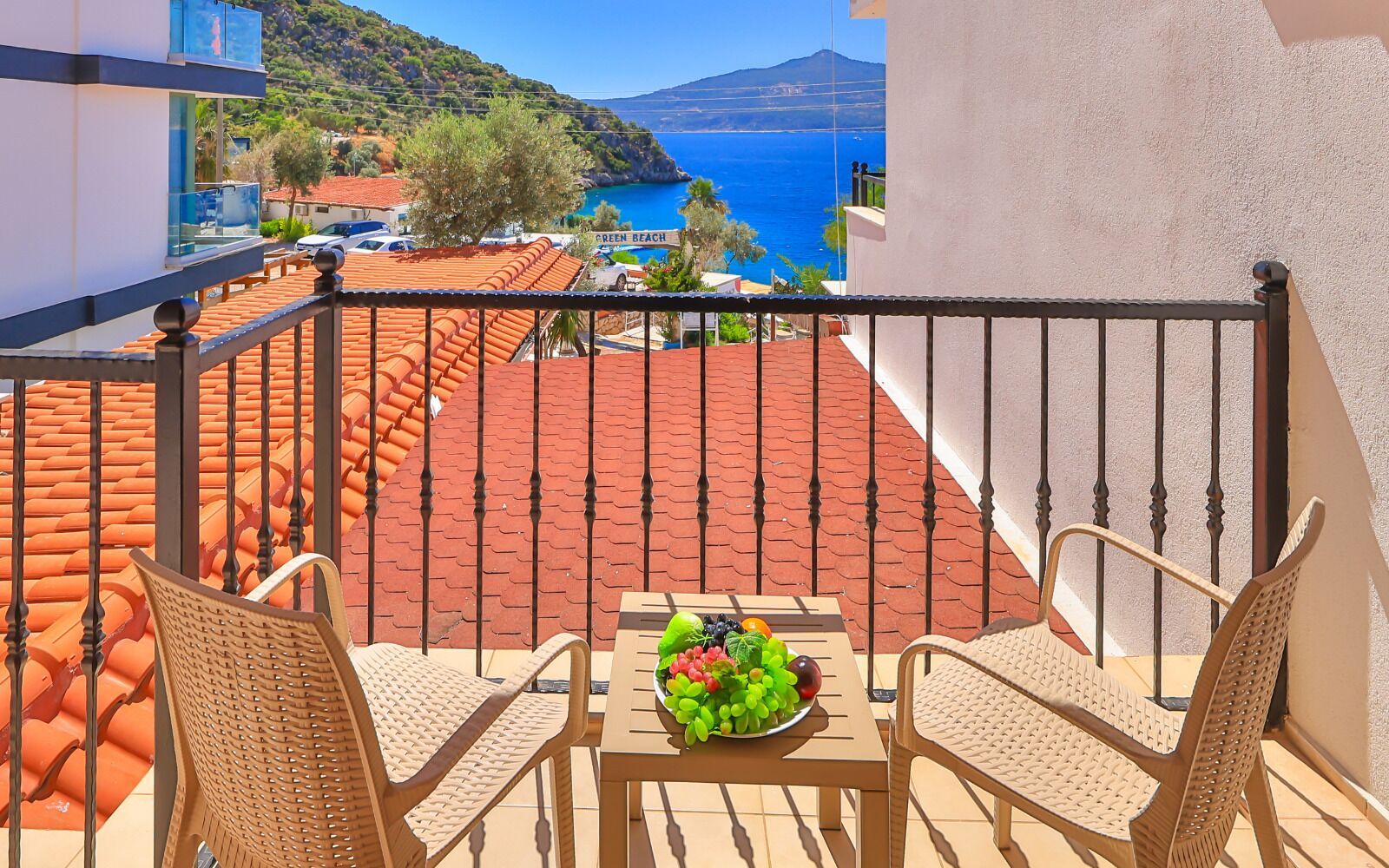 A balcony with wicker chairs, a table with a fruit bowl, and a view of the sea and a mountain.