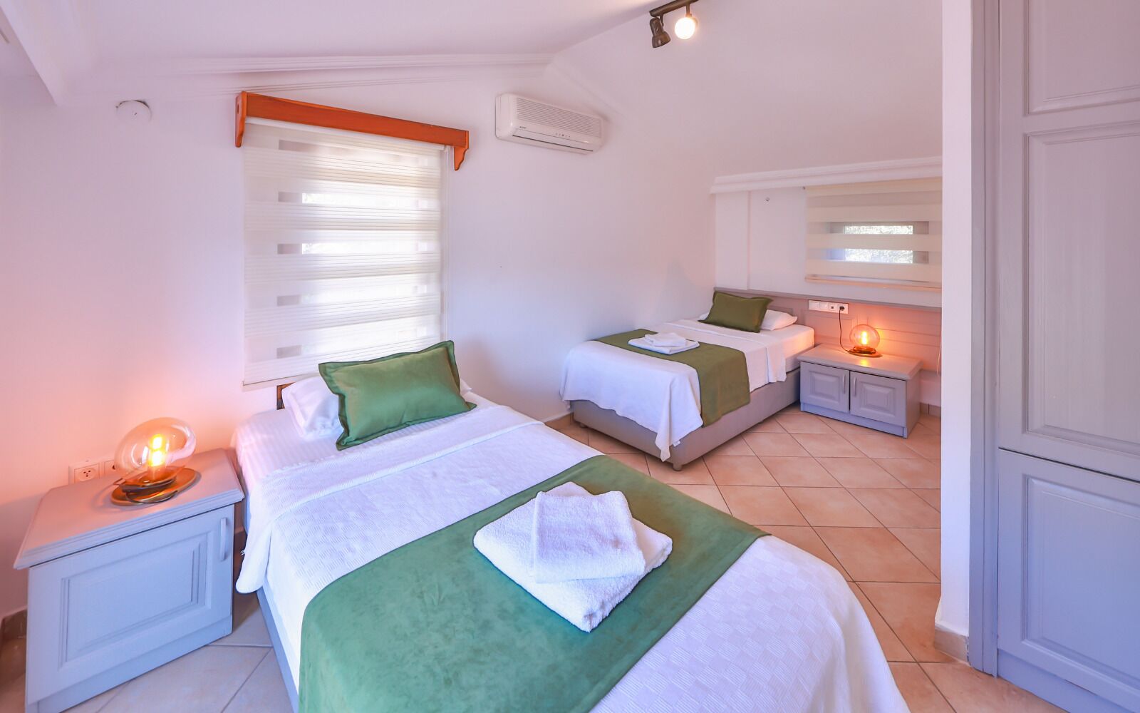 A hotel room with two beds, a bedside table with a lamp, a window with blinds, an air conditioning unit, and a nightstand with a lamp.