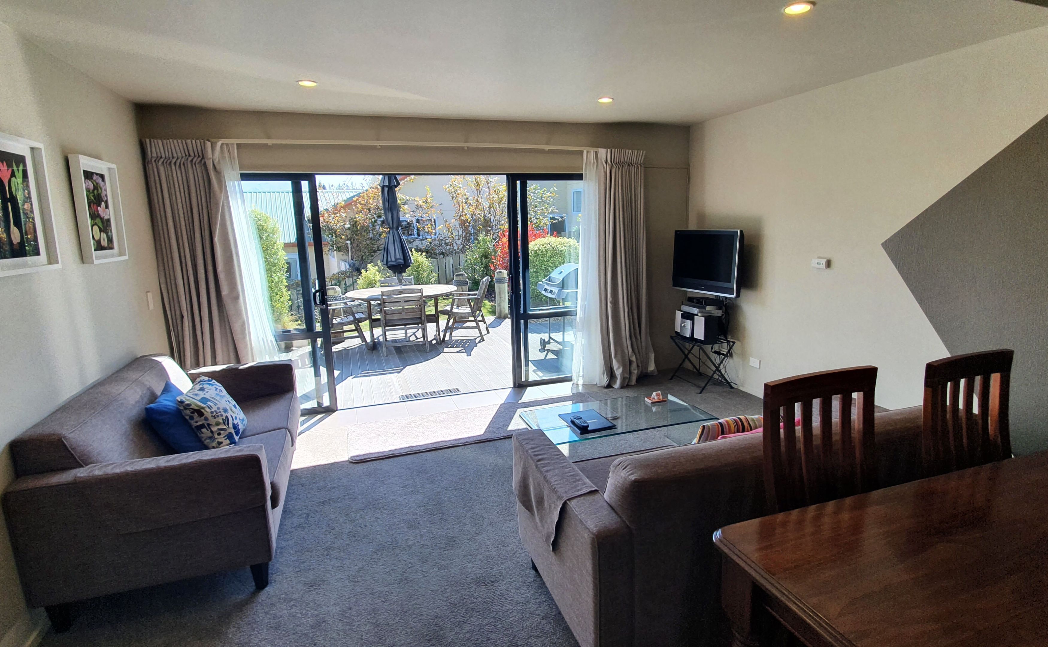 A living room with a sofa, a coffee table, and a TV. There is a sliding glass door leading to an outdoor patio with a table and chairs.