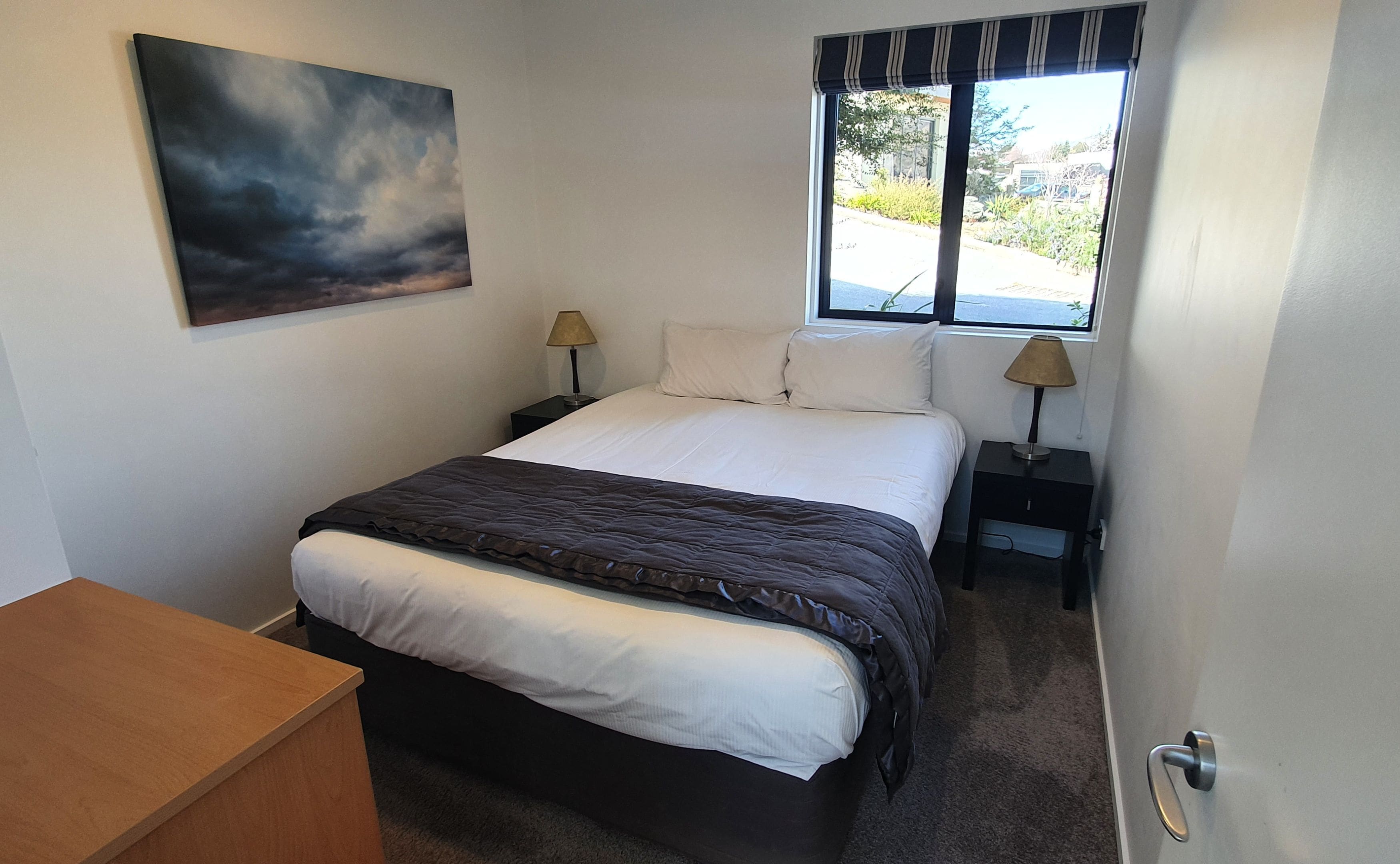 Un dormitorio con una cama, dos mesitas de noche, una ventana con cortinas, un cuadro en la pared y un escritorio de madera.
