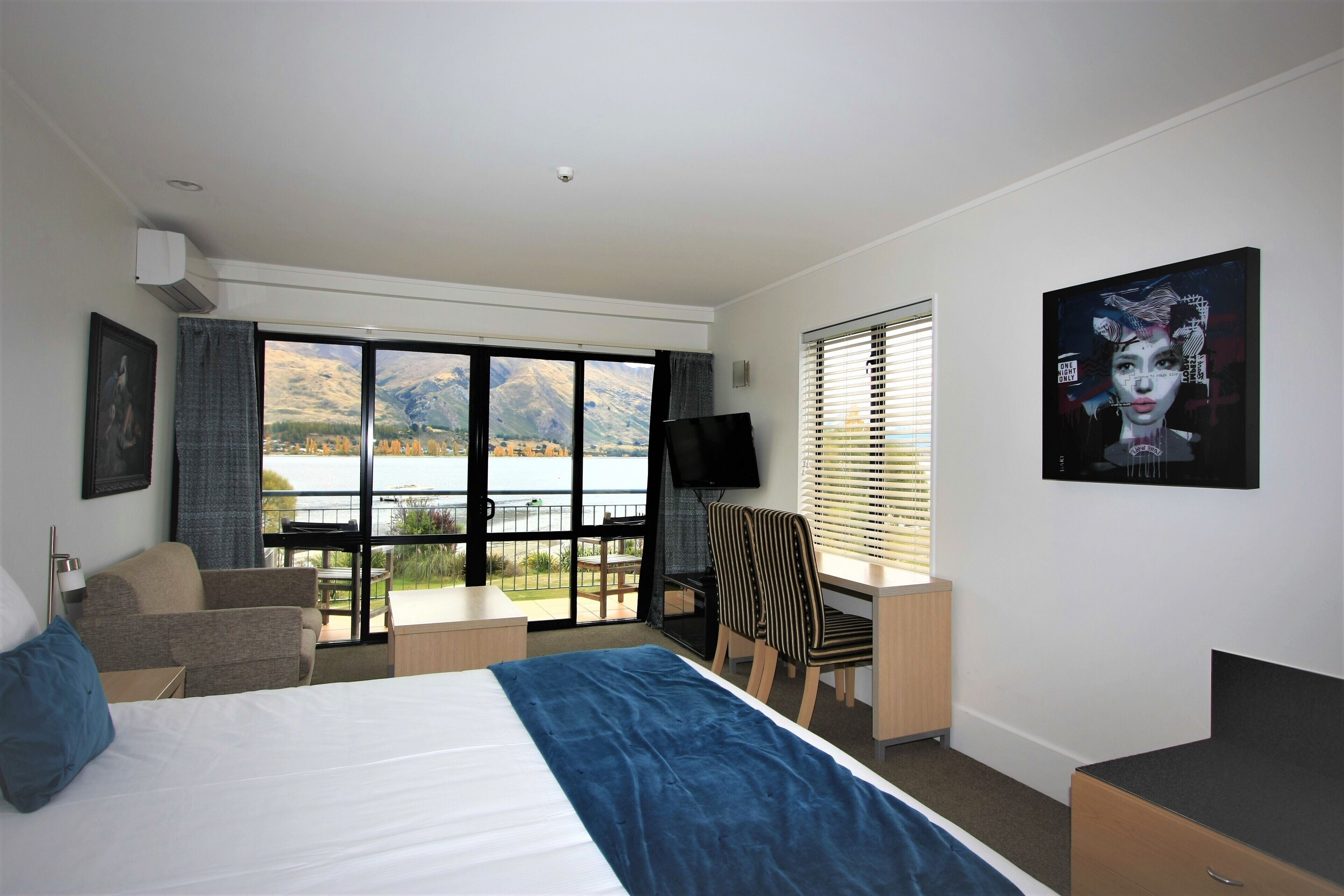 Quarto de hotel com uma cama grande, uma escrivaninha, uma cadeira e uma TV. Possui varanda com vista para montanhas e um lago.