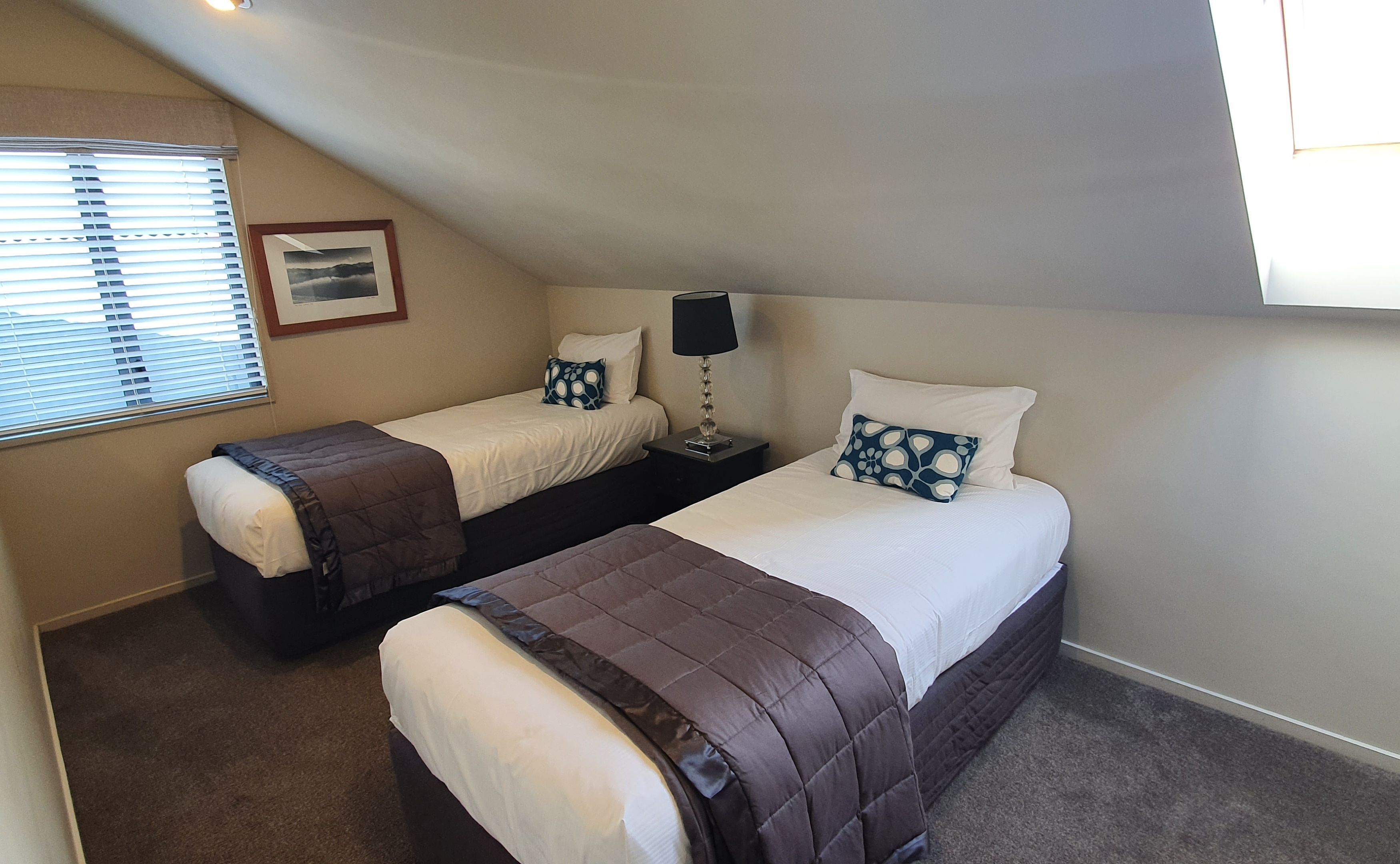 A bedroom with two single beds, a nightstand with a lamp, a framed picture on the wall, and a window with blinds.