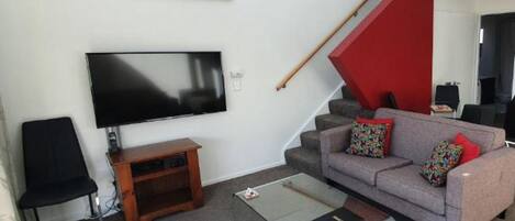 Un salon moderne doté d’un téléviseur à écran plat, d’un canapé agrémenté de coussins colorés, d’une table basse en verre et d’un escalier dont une partie est peinte en rouge.