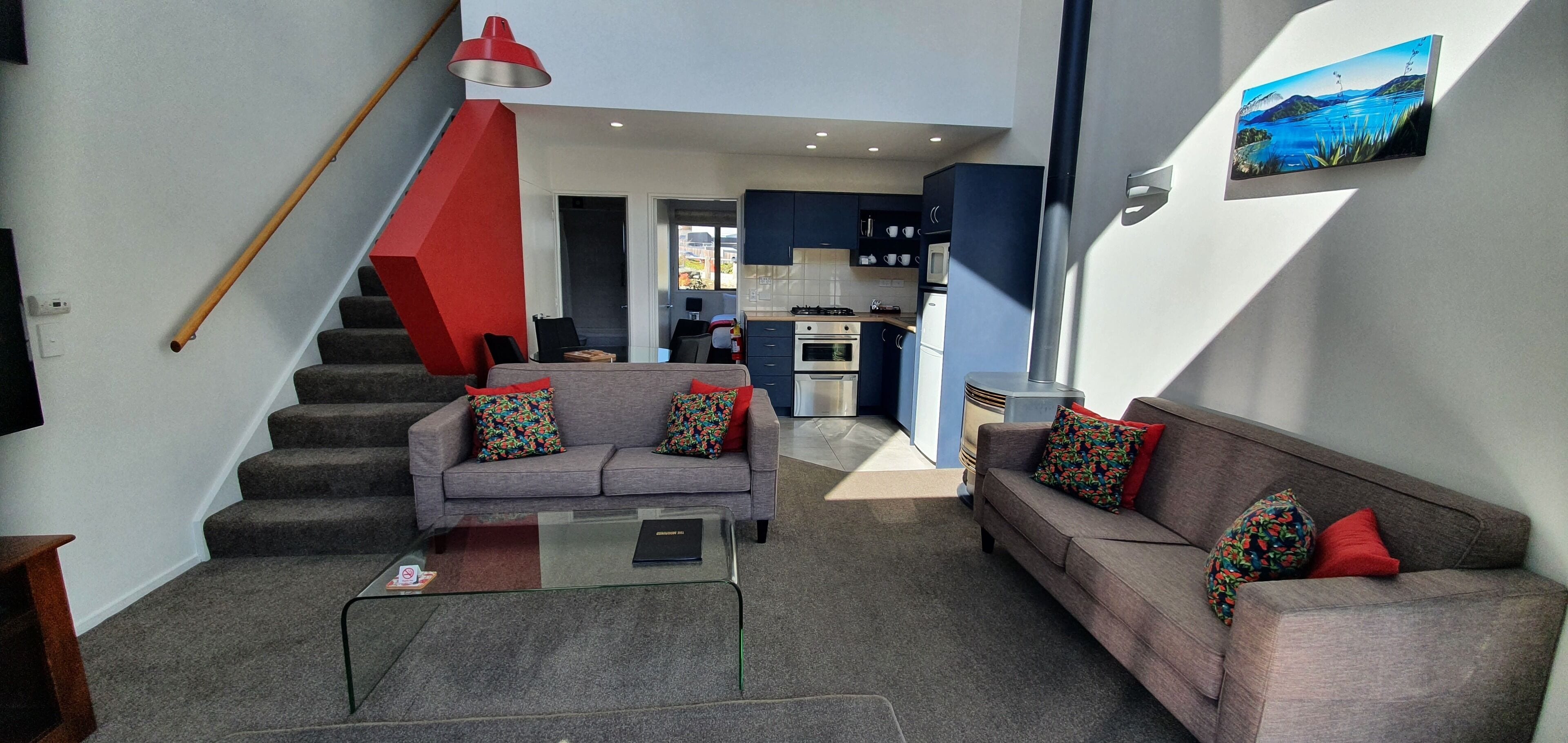 A modern living room with a staircase, a sofa set, a glass coffee table, and a kitchen in the background.
