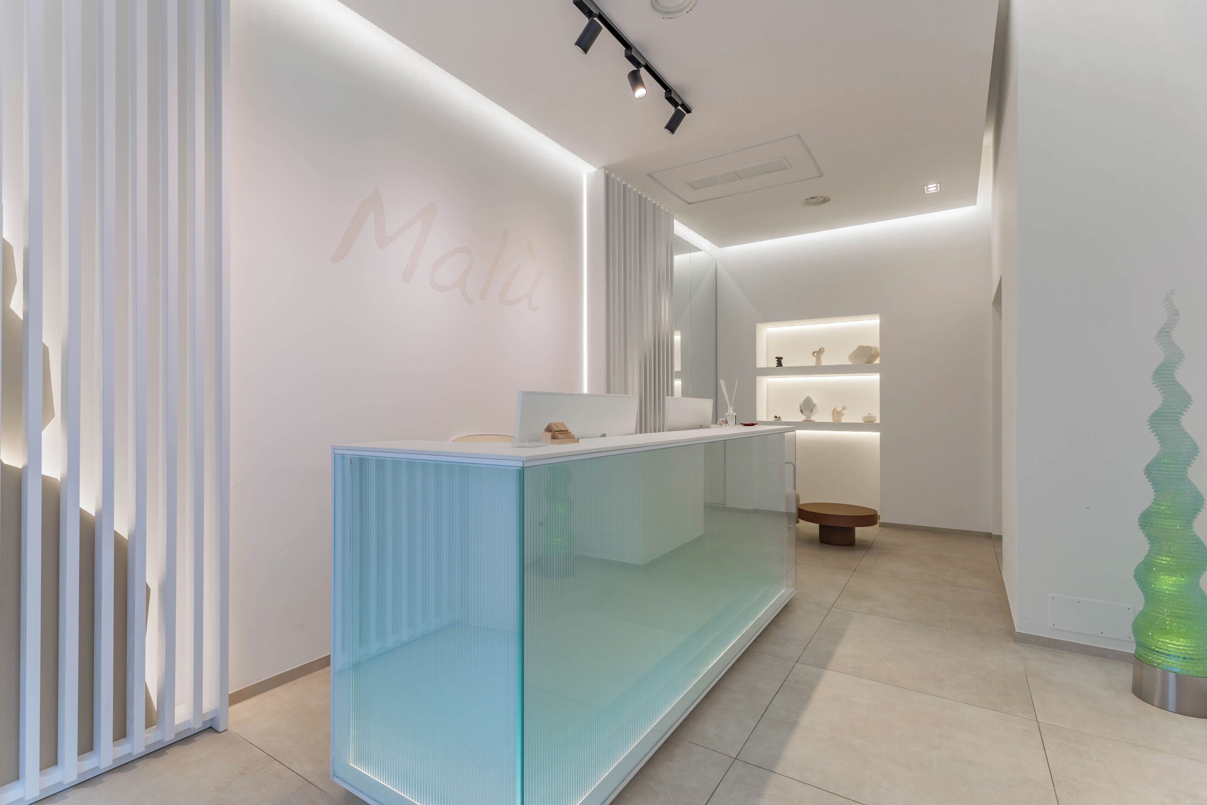 Un espace d’accueil avec un comptoir en verre, un mur sur lequel on peut apercevoir le mot « Malu » en partie, et une sculpture décorative en verre.